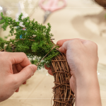 先以鐵絲穿過藤圈並收好，抓一把雪松，繞過後拉緊，再取一把雪松，在第一把根部接上，以此將雪松綁完。